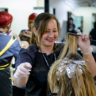 A hair stylist laughs with her client as she as she draws up a strand foiled hair with a comb